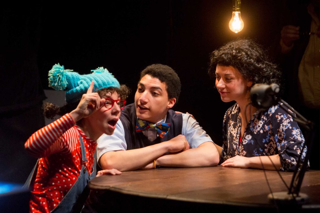 Three people sit on stage around a table on a stage, making expressive faces.