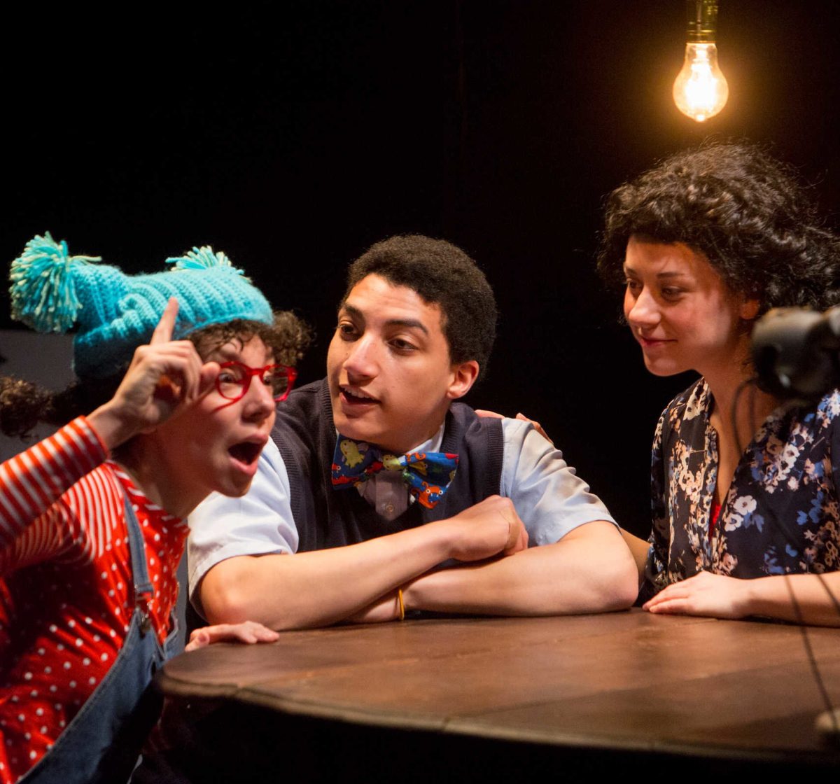Three people sit on stage around a table on a stage, making expressive faces.