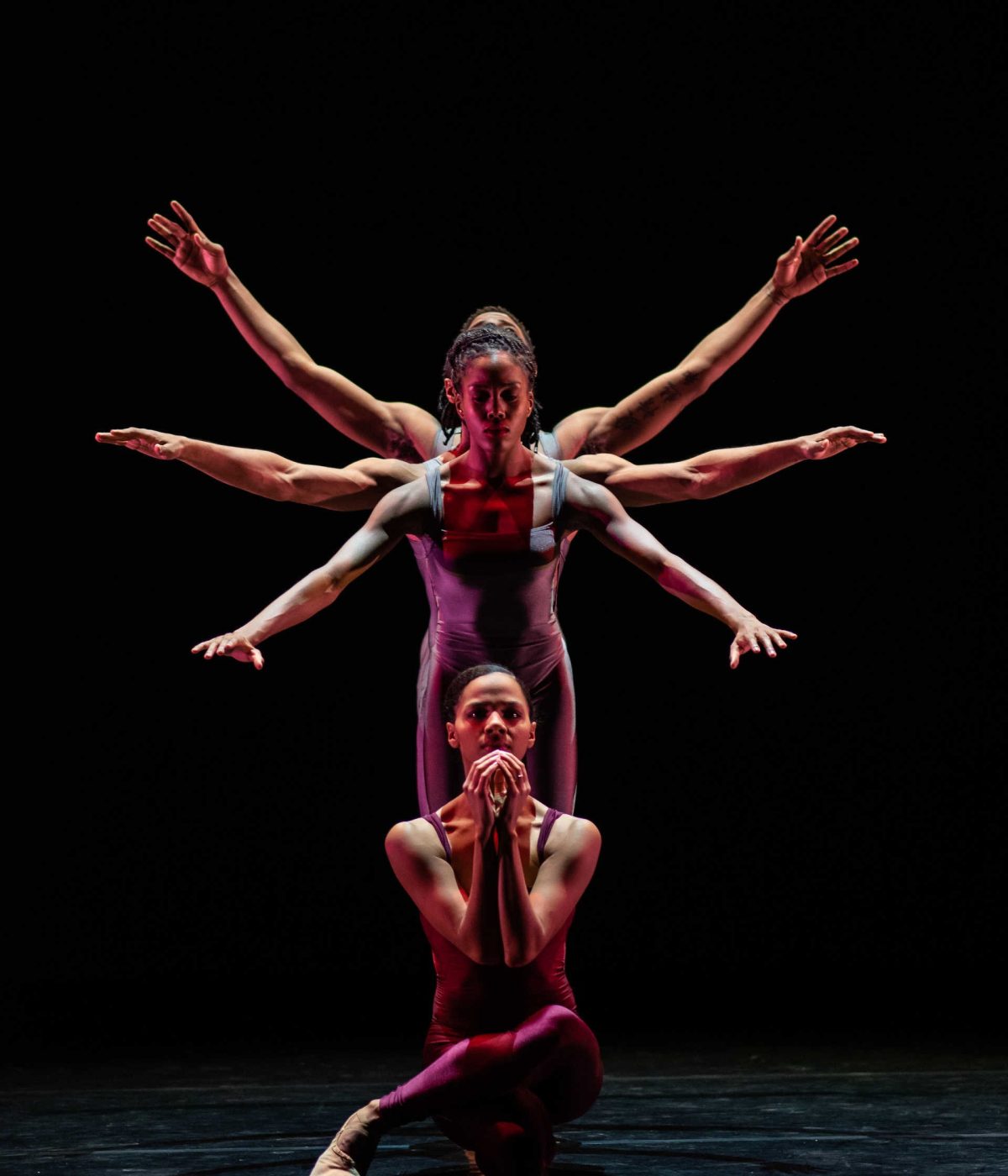 Four dancers on stage, standing one in front of the other, with their arms raised beside them at three different heights