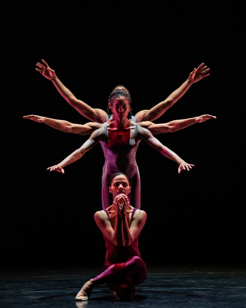 Four dancers on stage, standing one in front of the other, with their arms raised beside them at three different heights